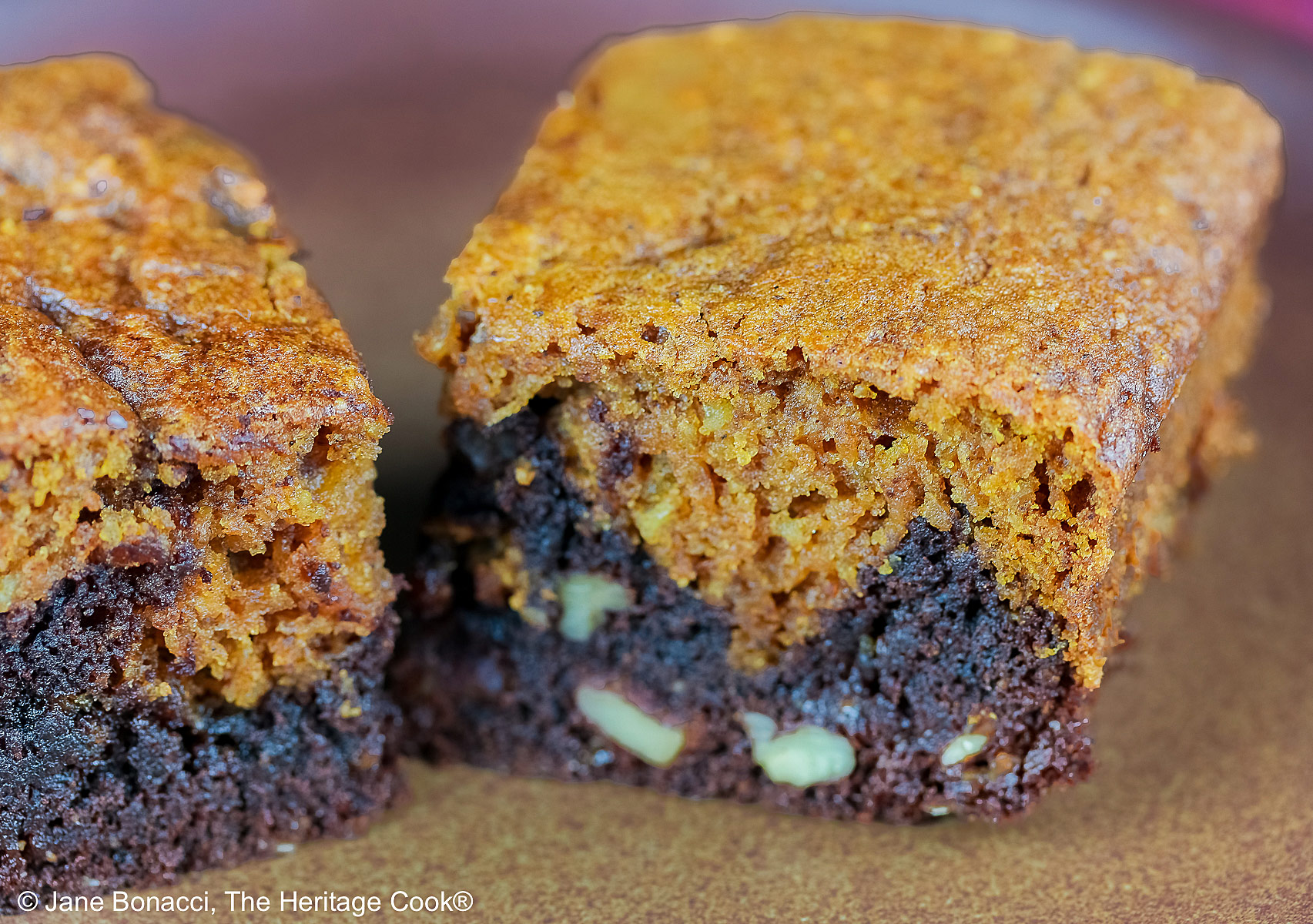 Pumpkin Swirl Brownies cut into squares and sitting on a dark plate, some stacked 3 high with a dark background that highlights the contrast between the dark brownies on the bottom with a topping of golden pumpkin cake on each piece © 2026 Jane Bonacci, The Heritage Cook.