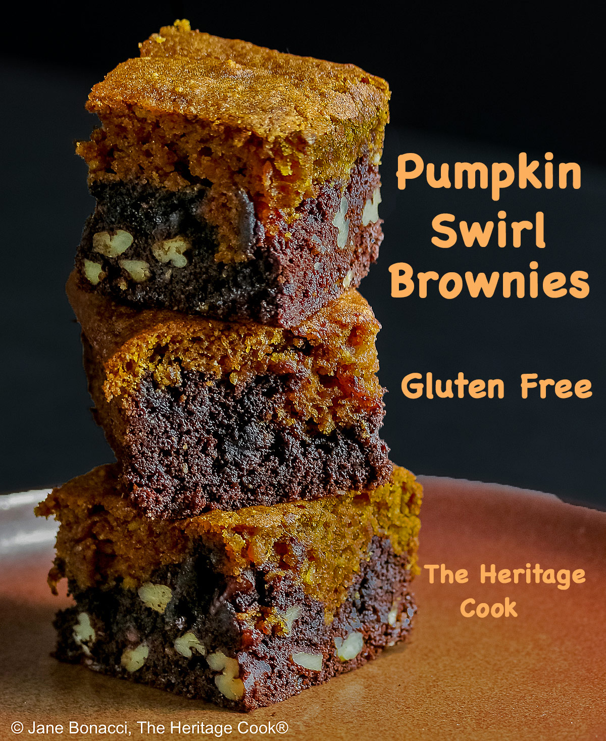 Pumpkin Swirl Brownies cut into squares and sitting on a dark plate, some stacked 3 high with a dark background that highlights the contrast between the dark brownies on the bottom with a topping of golden pumpkin cake on each piece © 2026 Jane Bonacci, The Heritage Cook.