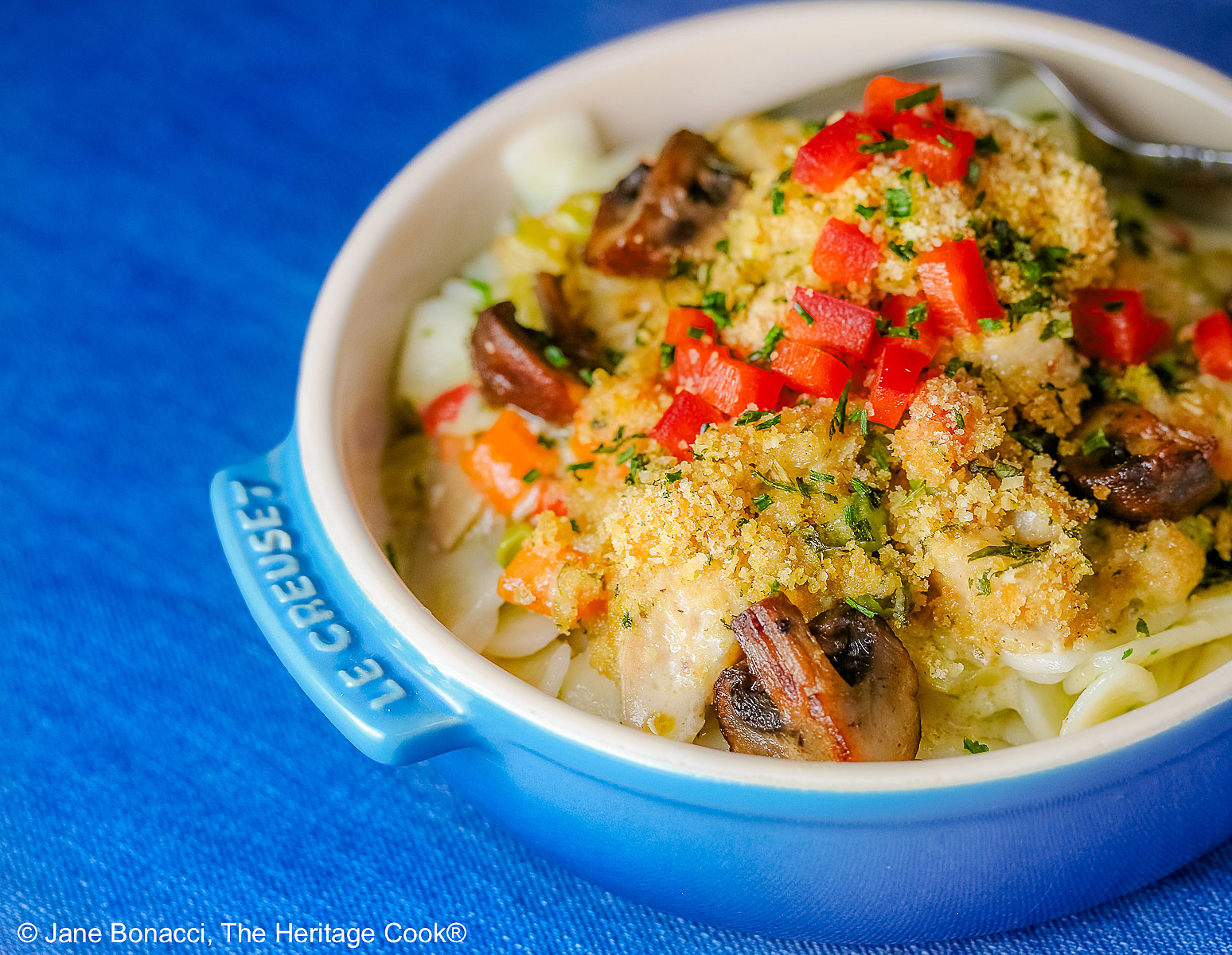 A blue round baking dish with handles on a vivid blue background, most with a bright multi-colored cloth beneath, some with a fork on the side, some with a white square plate beneath the bowl © 2026 Jane Bonacci, The Heritage Cook.
