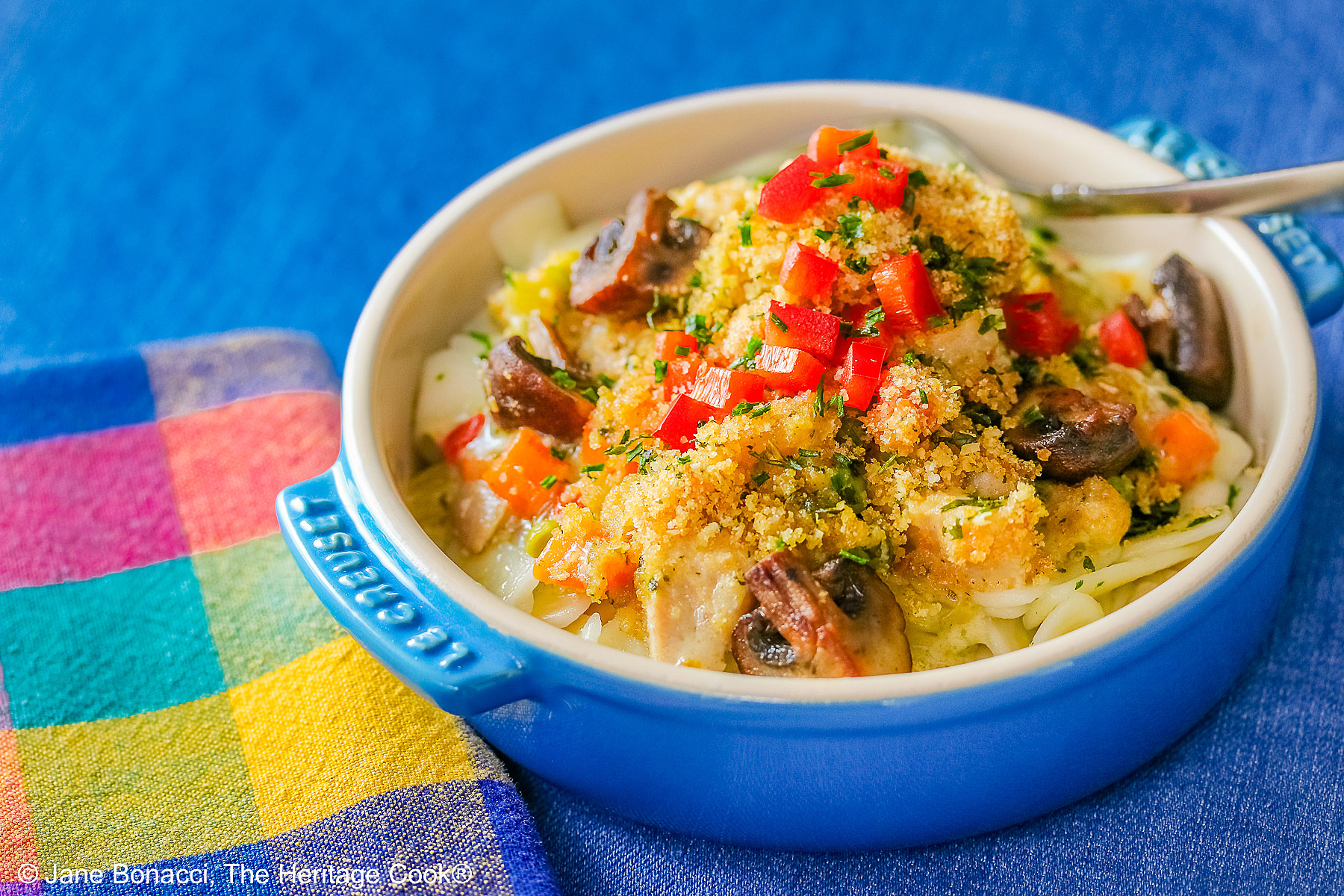 A blue round baking dish with handles on a vivid blue background, most with a bright multi-colored cloth beneath, some with a fork on the side, some with a white square plate beneath the bowl © 2026 Jane Bonacci, The Heritage Cook.