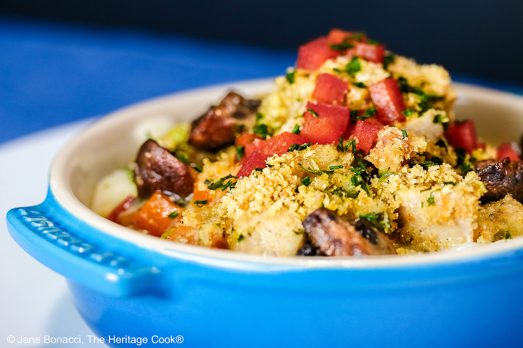 A blue round baking dish with handles on a vivid blue background, most with a bright multi-colored cloth beneath, some with a fork on the side, some with a white square plate beneath the bowl © 2026 Jane Bonacci, The Heritage Cook.