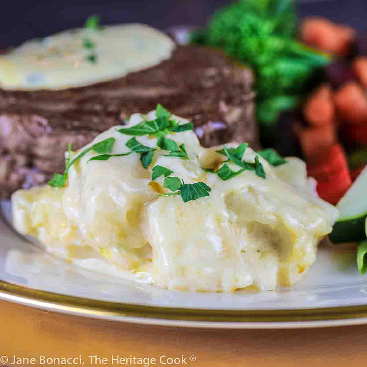 Scoop of Potatoes au Gratin sprinkled with chopped parsley on a gold rimmed plate, part of a festive meal with steak and brightly colored vegetables © 2023 Jane Bonacci, The Heritage Cook.