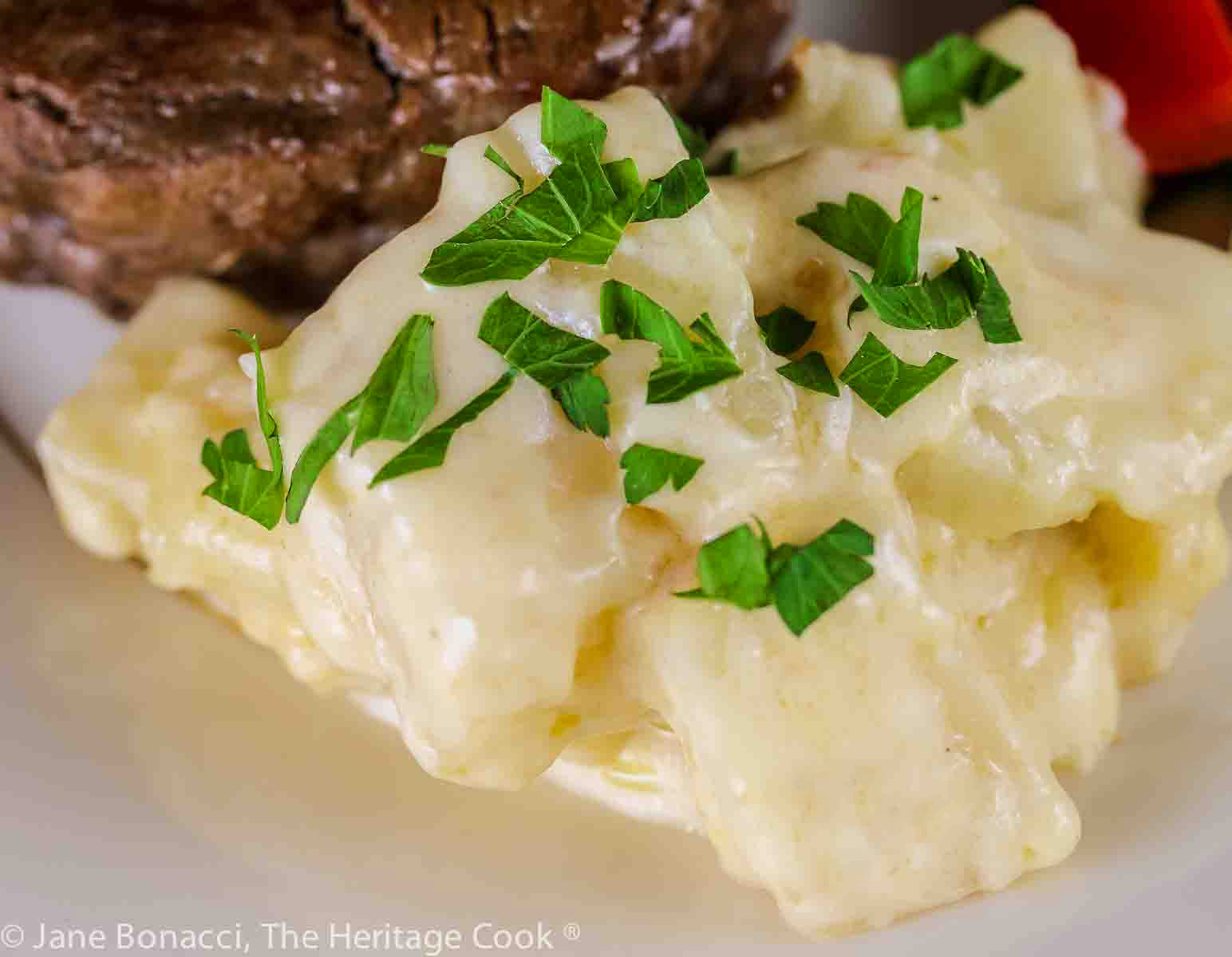 Scoop of Potatoes au Gratin sprinkled with chopped parsley on a gold rimmed plate, part of a festive meal with steak and brightly colored vegetables © 2023 Jane Bonacci, The Heritage Cook.