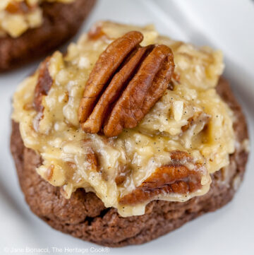 Three German Chocolate Cookies, chocolate cookies with coconut-pecan frosting on top, on a white rectangular platter with a cup of coffee behind on a black background © 2025 Jane Bonacci, The Heritage Cook.