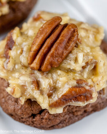 Three German Chocolate Cookies, chocolate cookies with coconut-pecan frosting on top, on a white rectangular platter with a cup of coffee behind on a black background © 2025 Jane Bonacci, The Heritage Cook.