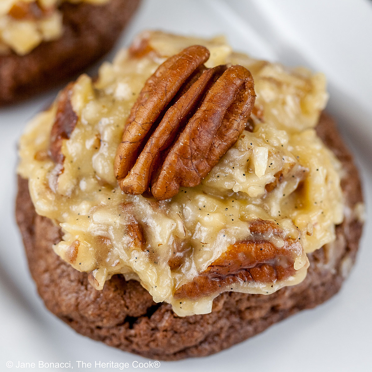 Three German Chocolate Cookies, chocolate cookies with coconut-pecan frosting on top, on a white rectangular platter with a cup of coffee behind on a black background © 2025 Jane Bonacci, The Heritage Cook.