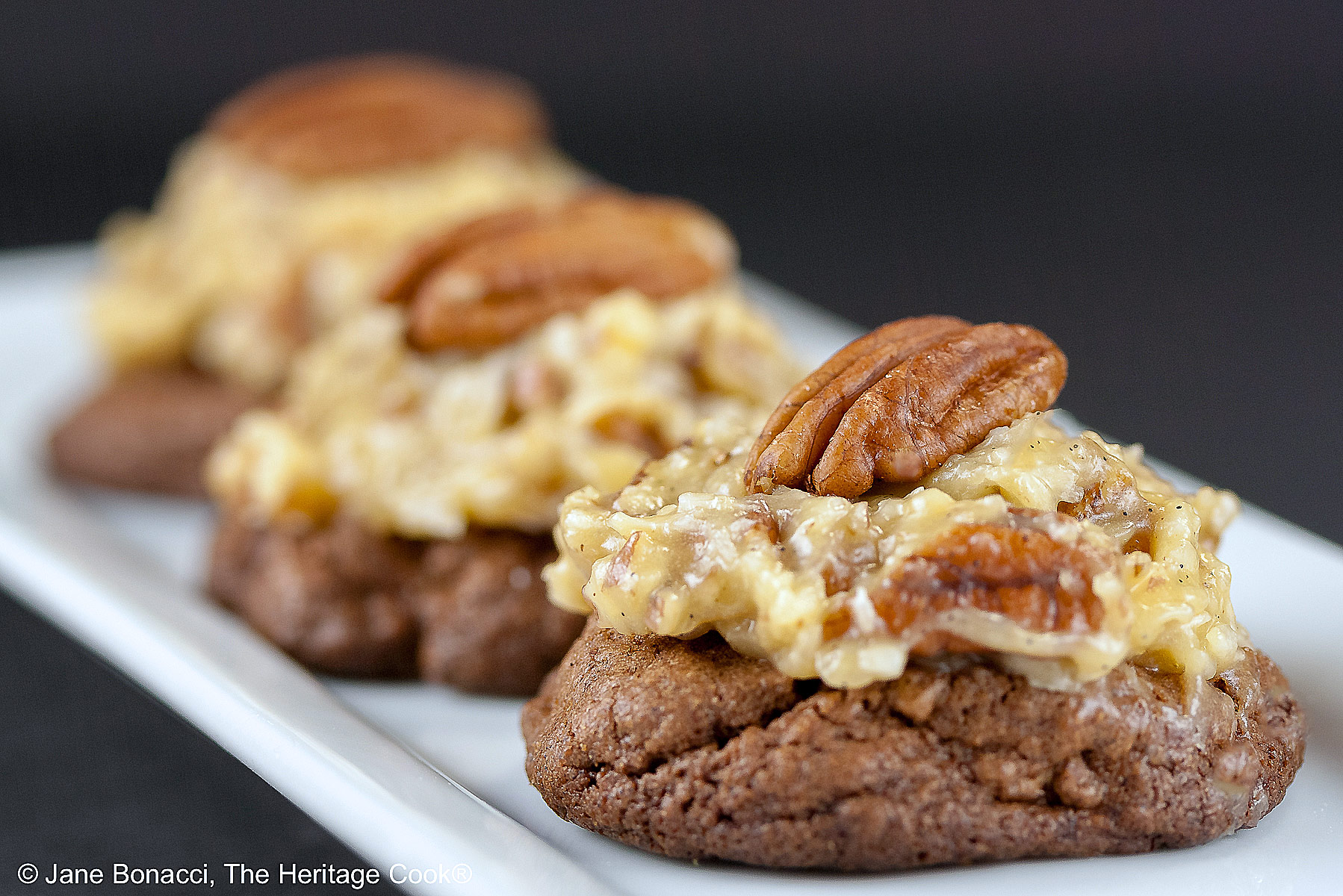 Three German Chocolate Cookies, chocolate cookies with coconut-pecan frosting on top, on a white rectangular platter with a cup of coffee behind on a black background © 2025 Jane Bonacci, The Heritage Cook.