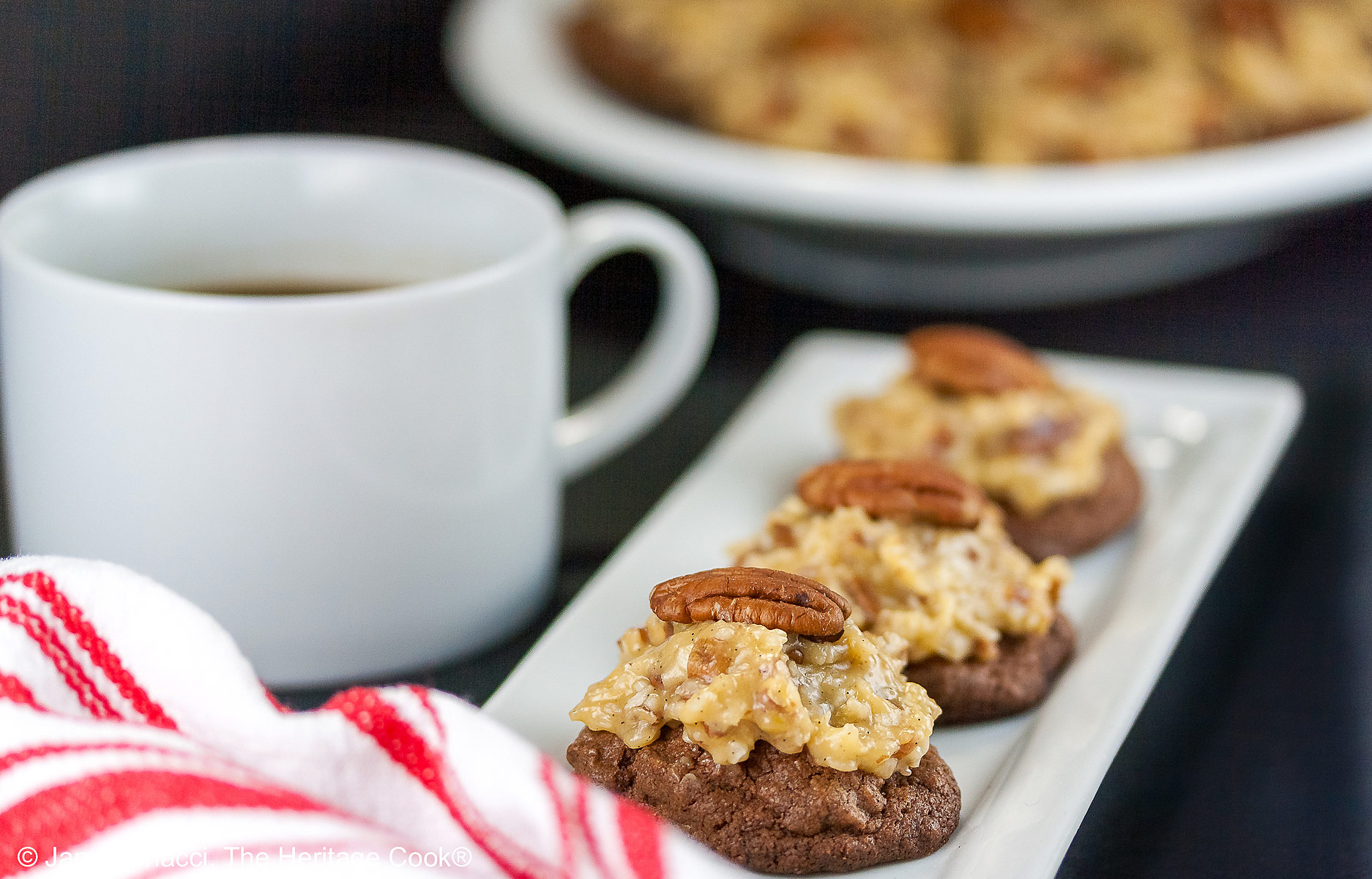 Three German Chocolate Cookies, chocolate cookies with coconut-pecan frosting on top, on a white rectangular platter with a cup of coffee behind on a black background © 2025 Jane Bonacci, The Heritage Cook.