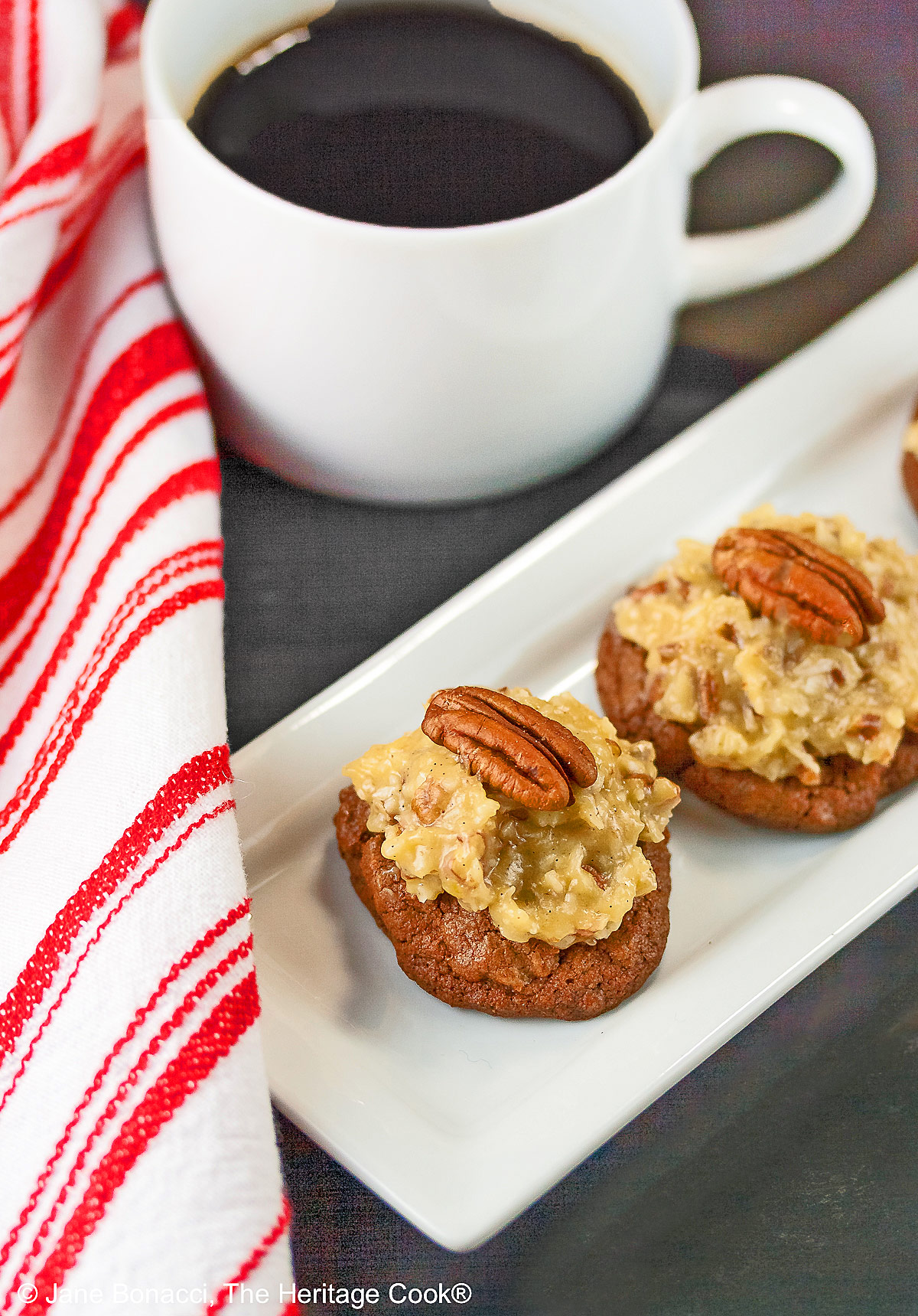Three German Chocolate Cookies, chocolate cookies with coconut-pecan frosting on top, on a white rectangular platter with a cup of coffee behind on a black background © 2025 Jane Bonacci, The Heritage Cook.