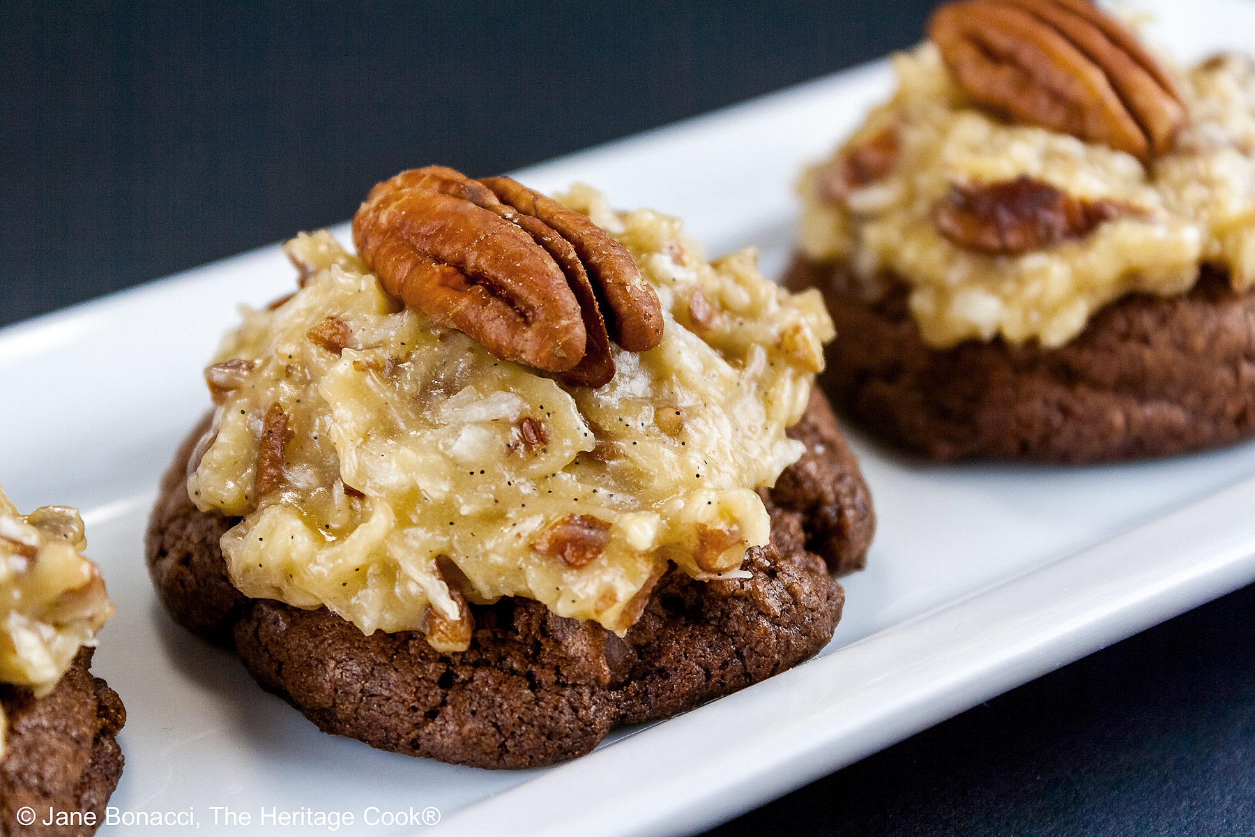 Three German Chocolate Cookies, chocolate cookies with coconut-pecan frosting on top, on a white rectangular platter with a cup of coffee behind on a black background © 2025 Jane Bonacci, The Heritage Cook.