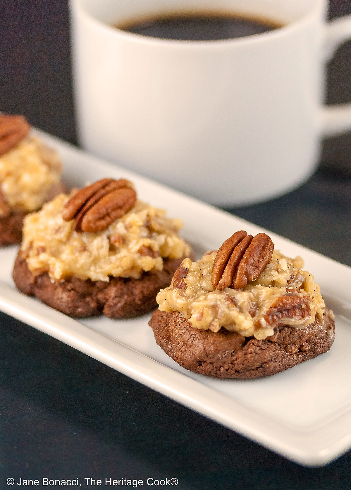 Three German Chocolate Cookies, chocolate cookies with coconut-pecan frosting on top, on a white rectangular platter with a cup of coffee behind on a black background © 2025 Jane Bonacci, The Heritage Cook.