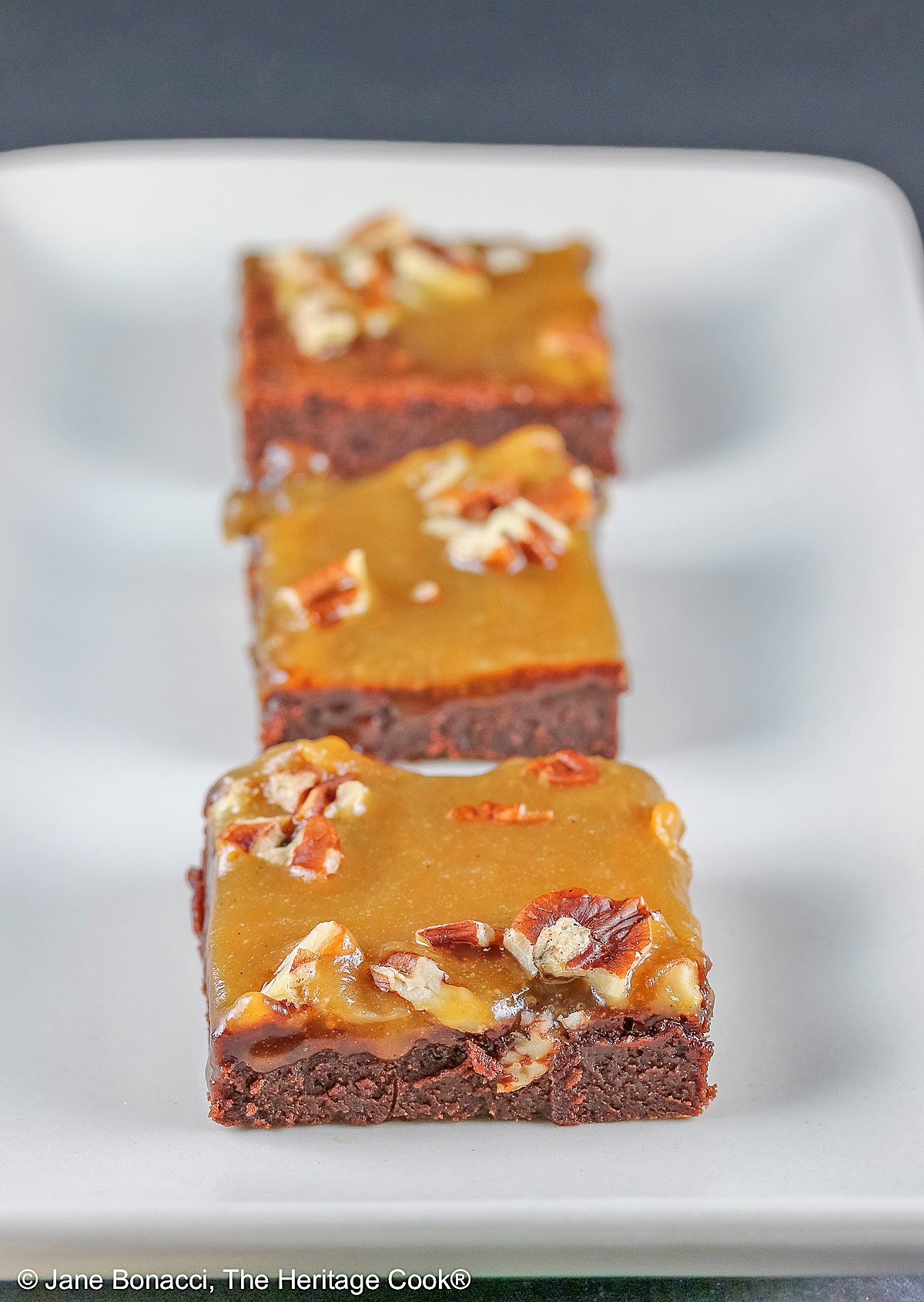 Line of 3 caramel coated brownies on a long rectangular white platter, sitting on a black background, close up of the brownies © 2026 Jane Bonacci, The Heritage Cook.