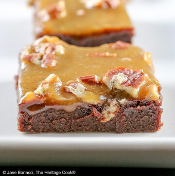 Line of 3 caramel coated brownies on a long rectangular white platter, sitting on a black background, close up of the brownies © 2026 Jane Bonacci, The Heritage Cook.