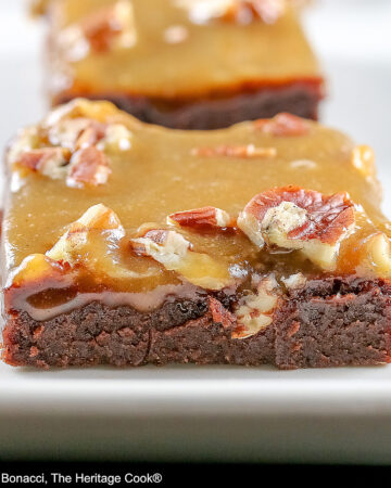 Line of 3 caramel coated brownies on a long rectangular white platter, sitting on a black background, close up of the brownies © 2026 Jane Bonacci, The Heritage Cook.