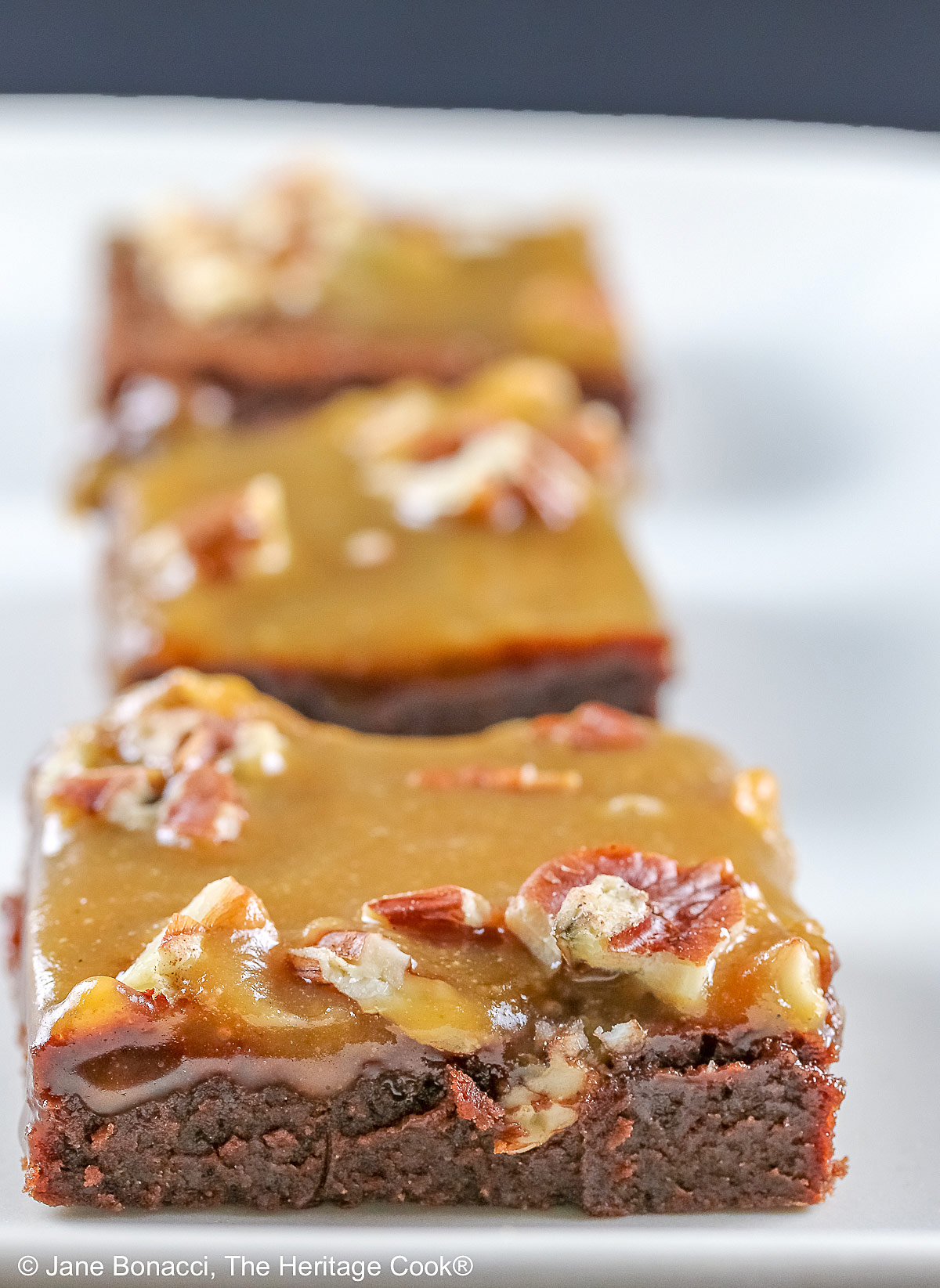 Line of 3 caramel coated brownies on a long rectangular white platter, sitting on a black background, close up of the brownies © 2026 Jane Bonacci, The Heritage Cook.