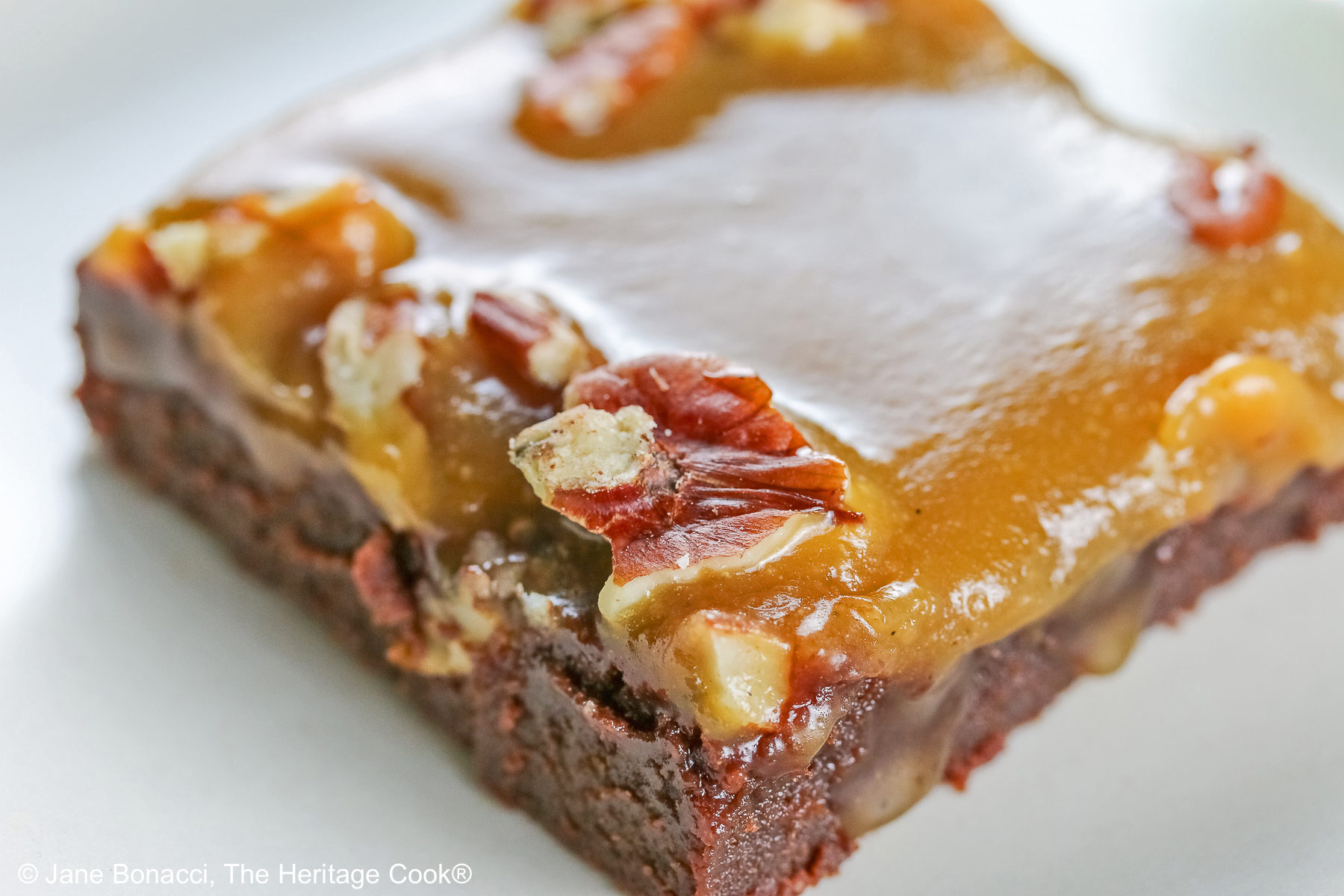 Line of 3 caramel coated brownies on a long rectangular white platter, sitting on a black background, close up of the brownies © 2026 Jane Bonacci, The Heritage Cook.