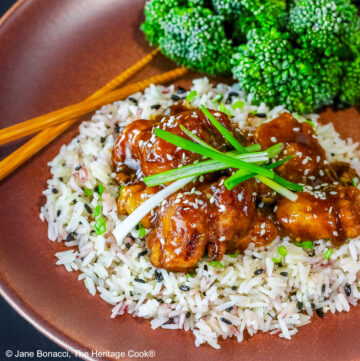 General Tso’s Chicken pieces on a bed of white rice with green onions and black sesame seeds, sprinkled with white sesame seeds, topped with slivered green onions, served with raw ginger and broccoli on a brick red plate © 2025 Jane Bonacci, The Heritage Cook.