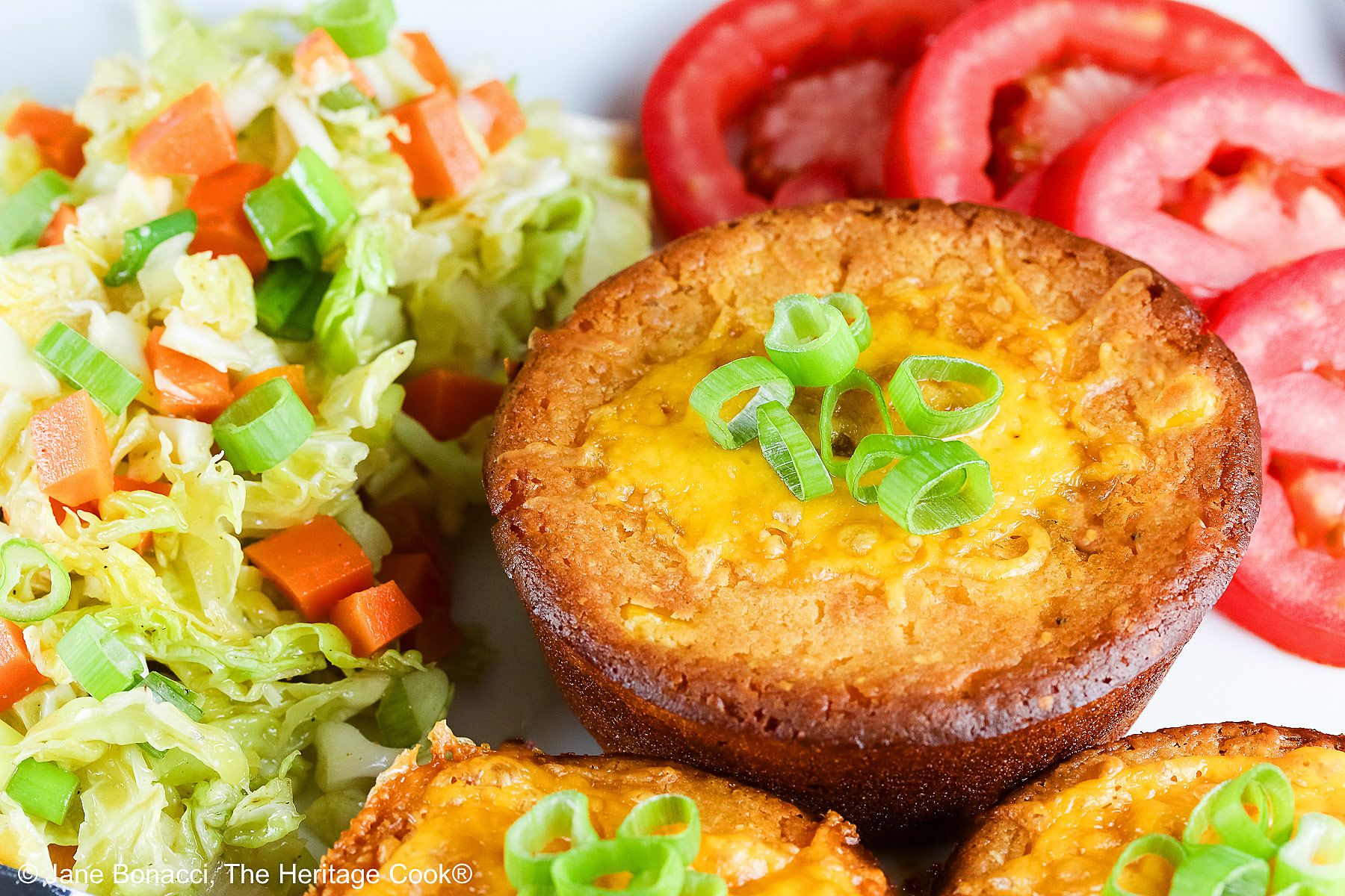 Cheesy Sloppy Joe Tamale Cups with sliced green onions on top on a white plate with colorful coleslaw and bright red tomato slices sitting on a red and white cloth © 2025 Jane Bonacci, The Heritage Cook.