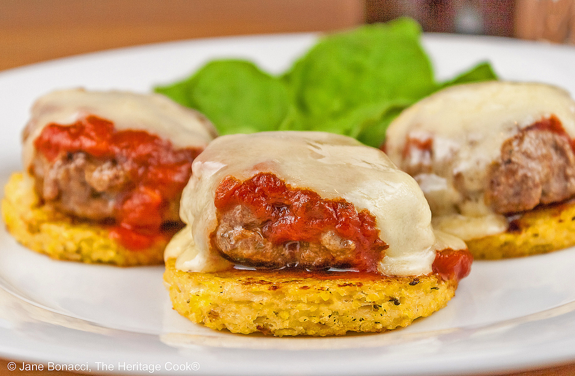 Three polenta disks topped with meatballs, melted cheese, and chopped basil on a white plate with a bunch of basil tucked in the back. There is a bottle of wine and filled glass behind the plate and everything is sitting on a pine wooden table © 2026 Jane Bonacci, The Heritage Cook.