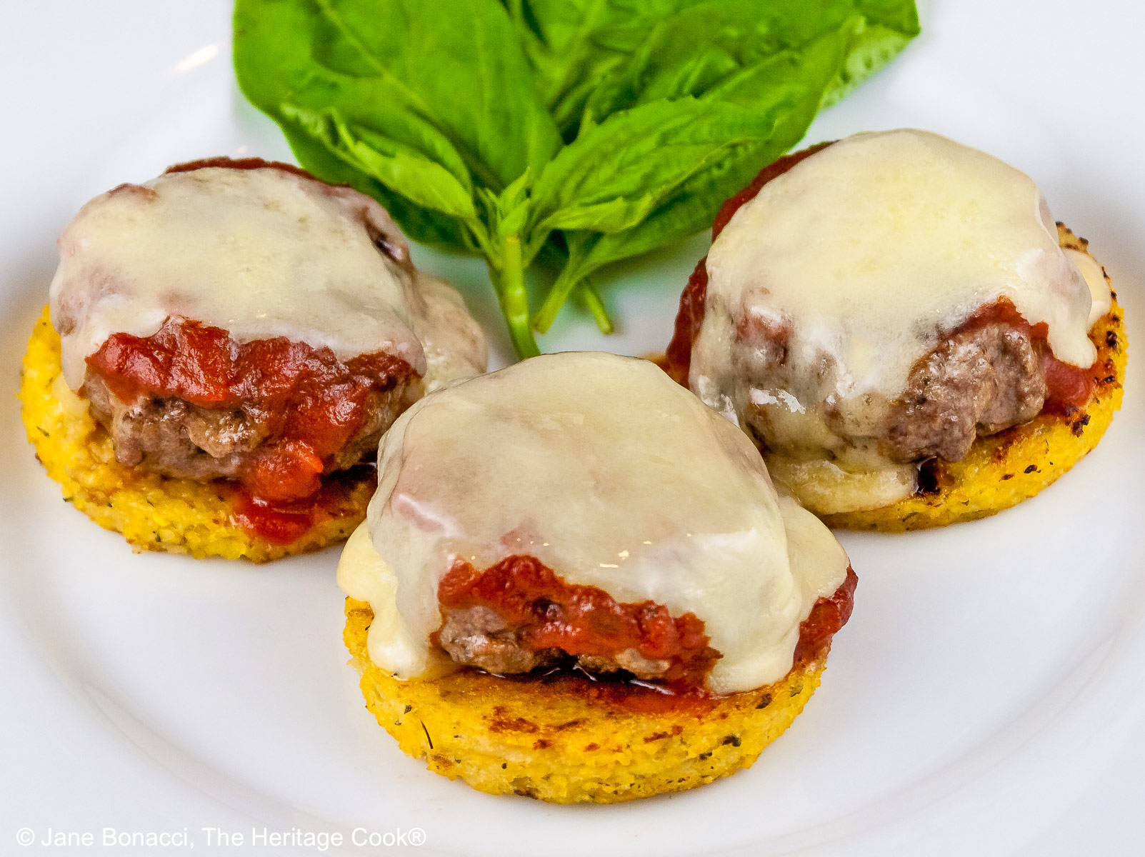 Three polenta disks topped with meatballs, melted cheese, and chopped basil on a white plate with a bunch of basil tucked in the back. There is a bottle of wine and filled glass behind the plate and everything is sitting on a pine wooden table © 2026 Jane Bonacci, The Heritage Cook.