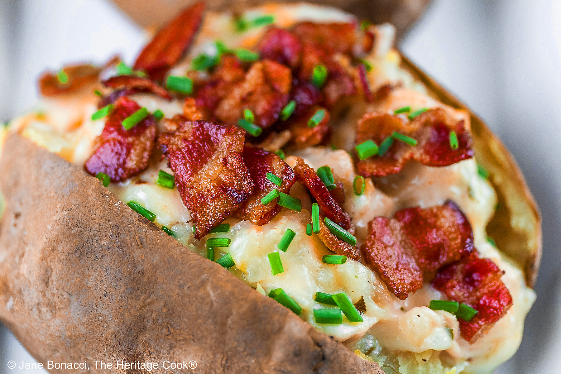 Baked potatoes split lengthwise and stuffed with a creamy cheese sauce with chicken, sitting on a white rectangle platter with a black background, topped with chopped bacon and chopped chives © 2025 Jane Bonacci, The Heritage Cook.
