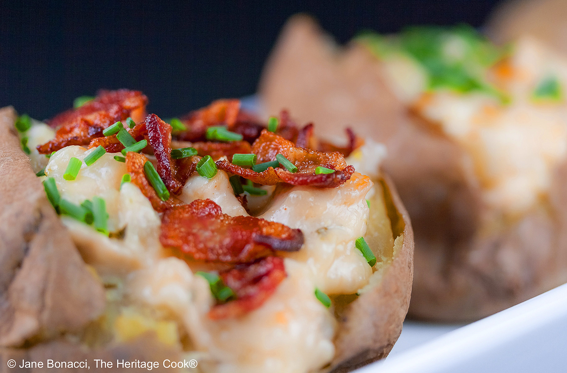 Baked potatoes split lengthwise and stuffed with a creamy cheese sauce with chicken, sitting on a white rectangle platter with a black background, topped with chopped bacon and chopped chives © 2025 Jane Bonacci, The Heritage Cook.