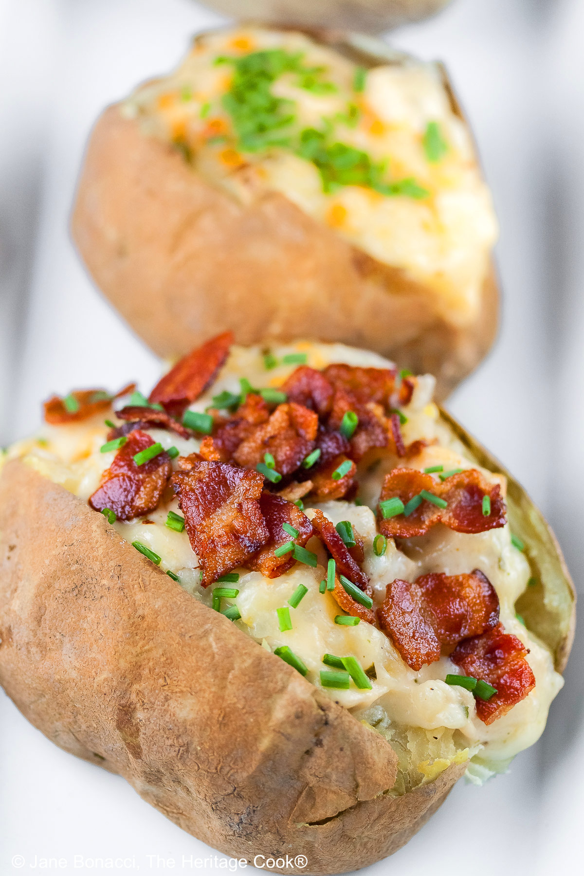 Baked potatoes split lengthwise and stuffed with a creamy cheese sauce with chicken, sitting on a white rectangle platter with a black background, topped with chopped bacon and chopped chives © 2025 Jane Bonacci, The Heritage Cook.