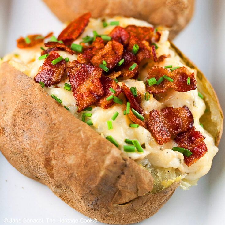 Baked potatoes split lengthwise and stuffed with a creamy cheese sauce with chicken, sitting on a white rectangle platter with a black background, topped with chopped bacon and chopped chives © 2025 Jane Bonacci, The Heritage Cook.