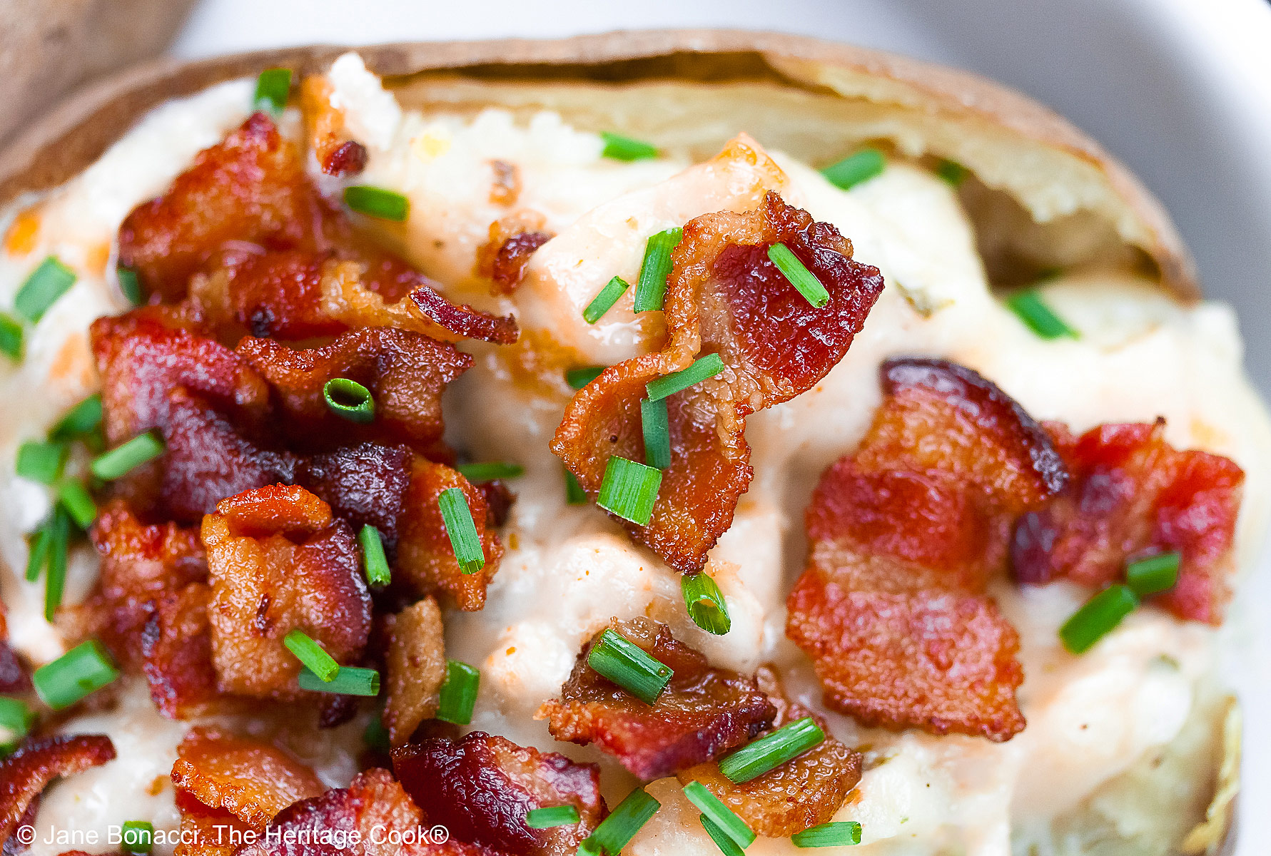 Baked potatoes split lengthwise and stuffed with a creamy cheese sauce with chicken, sitting on a white rectangle platter with a black background, topped with chopped bacon and chopped chives © 2025 Jane Bonacci, The Heritage Cook.