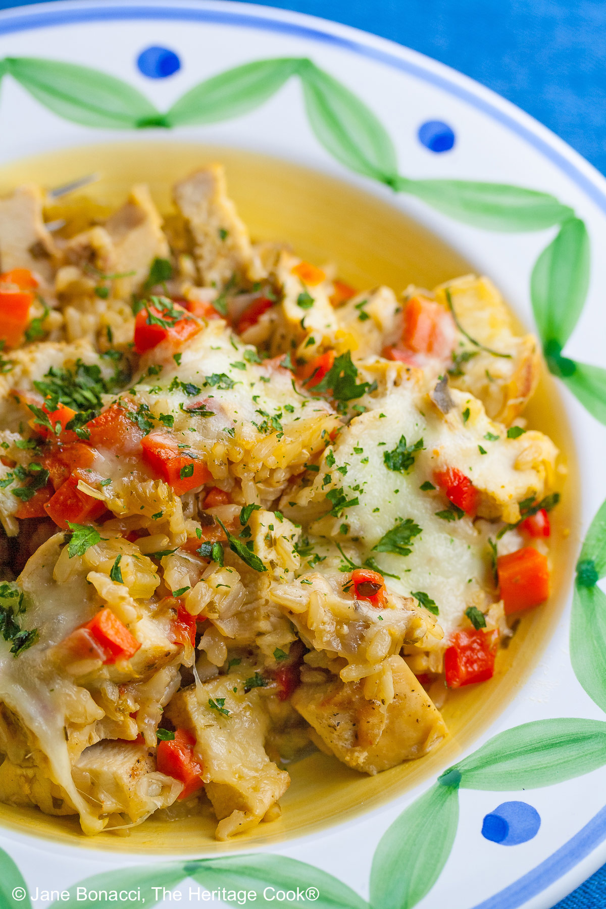 Golden rice mixed with red bell peppers, onions, and chicken, sprinkled with green parsley, this Oven Baked Chicken Risotto is in a bowl with a wide rim decorated with green and blue, sitting on a vivid blue background © 2026 Jane Bonacci, The Heritage Cook.