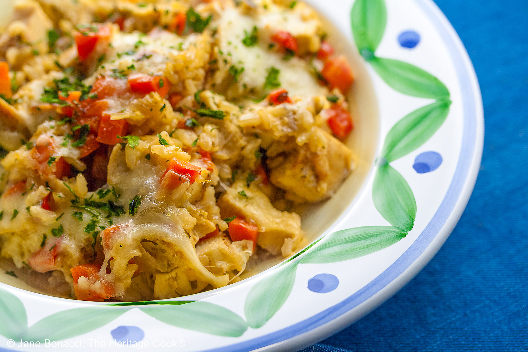 Golden rice mixed with red bell peppers, onions, and chicken, sprinkled with green parsley, this Oven Baked Chicken Risotto is in a bowl with a wide rim decorated with green and blue, sitting on a vivid blue background © 2026 Jane Bonacci, The Heritage Cook.