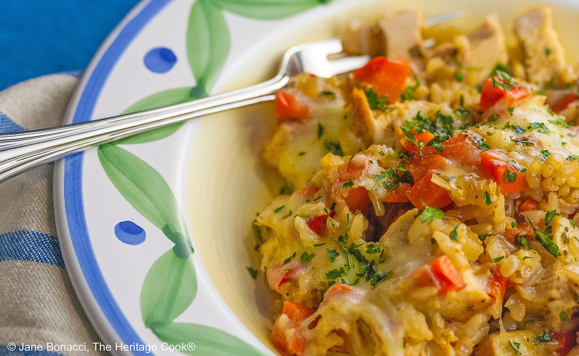 Golden rice mixed with red bell peppers, onions, and chicken, sprinkled with green parsley, this Oven Baked Chicken Risotto is in a bowl with a wide rim decorated with green and blue, sitting on a vivid blue background © 2026 Jane Bonacci, The Heritage Cook.