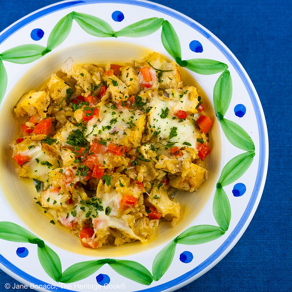 Golden rice mixed with red bell peppers, onions, and chicken, sprinkled with green parsley, this Oven Baked Chicken Risotto is in a bowl with a wide rim decorated with green and blue, sitting on a vivid blue background © 2026 Jane Bonacci, The Heritage Cook.