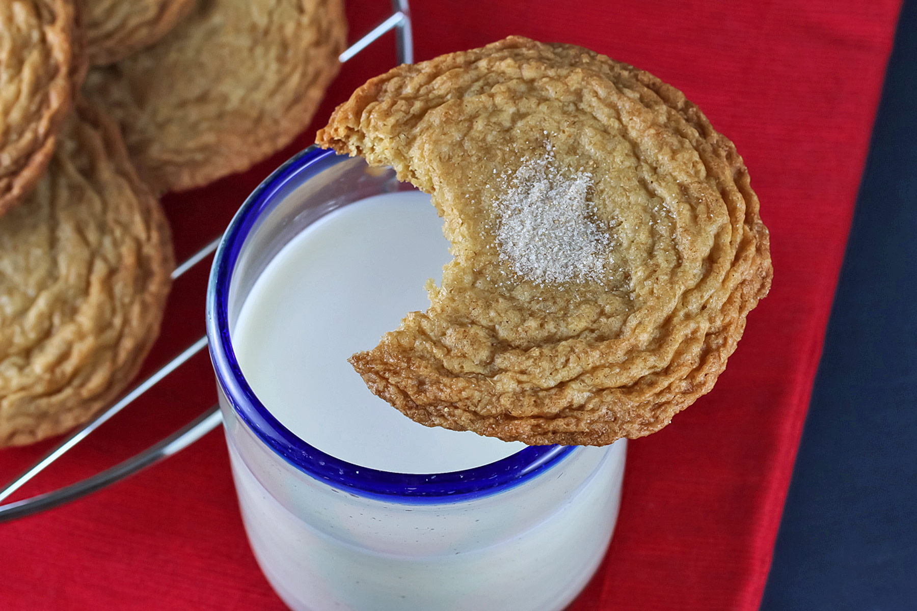 Stacks of thin Gingerdoodle Cookies with White Chocolate on a red plate placed on a dark blue cloth, some with a glass of milk in the background © 2024 Jane Bonacci, The Heritage Cook. 