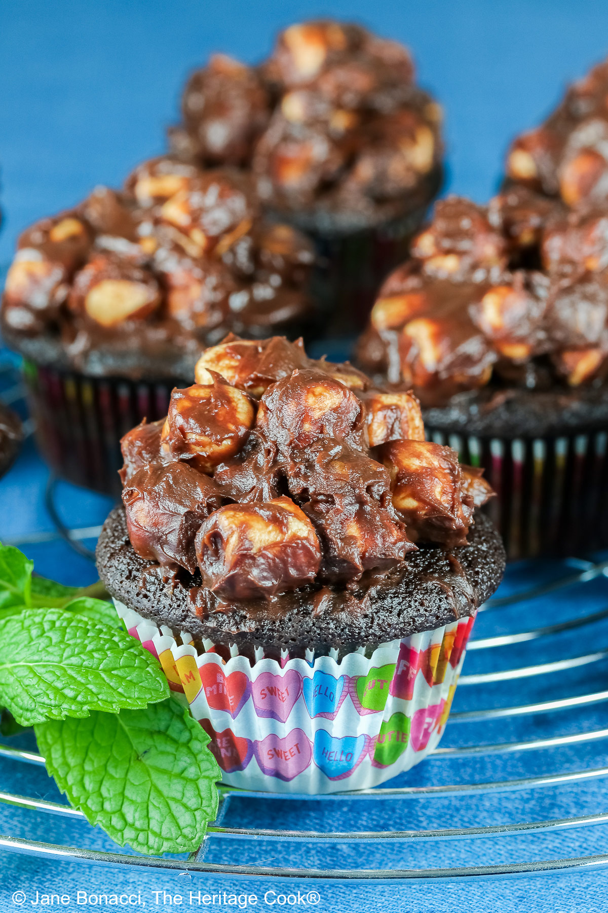 Rocky Road Chocolate Cupcakes in brightly patterned liners, sitting on a circular wire rack on top of a bright blue background and piled high with ganache covered chopped nuts and mini marshmallows © 2026 Jane Bonacci, The Heritage Cook.