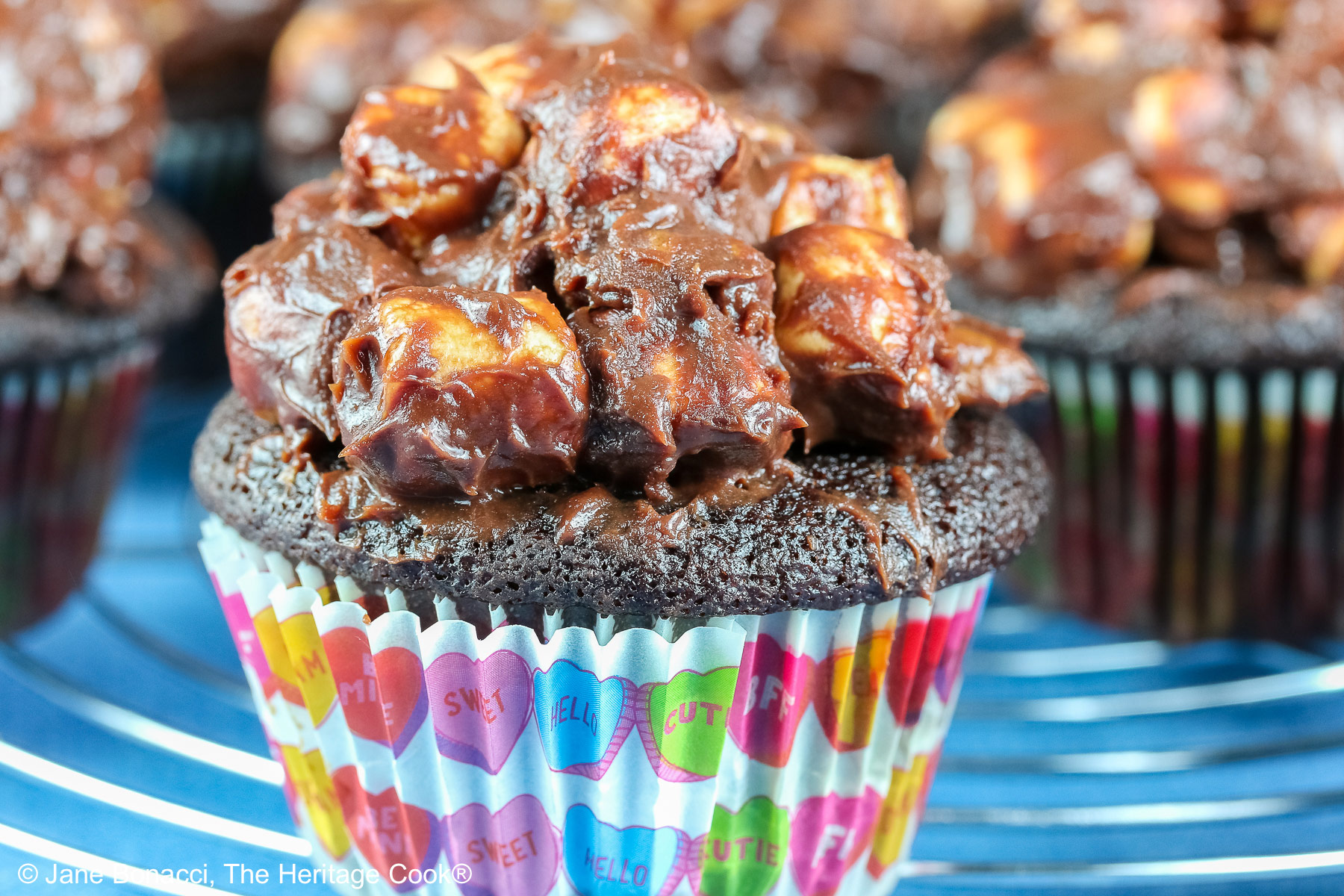 Rocky Road Chocolate Cupcakes in brightly patterned liners, sitting on a circular wire rack on top of a bright blue background and piled high with ganache covered chopped nuts and mini marshmallows © 2026 Jane Bonacci, The Heritage Cook.