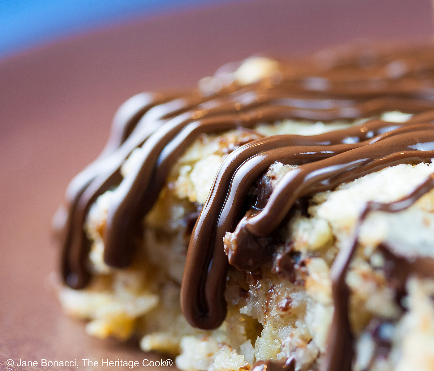 Close up of chocolate drizzle. Triangles and square of 7 Layer Congo Bars drizzled with chocolate sitting on a deep brown plate on top of a blue background © 2025 Jane Bonacci, The Heritage Cook. 