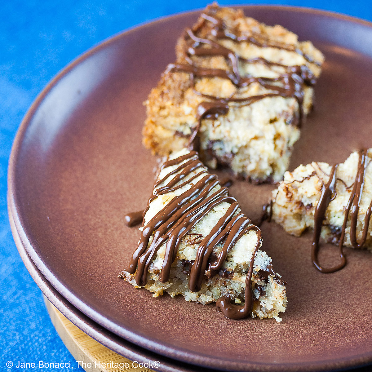 Triangles and square of 7 Layer Congo Bars drizzled with chocolate sitting on a deep brown plate on top of a blue background © 2025 Jane Bonacci, The Heritage Cook.