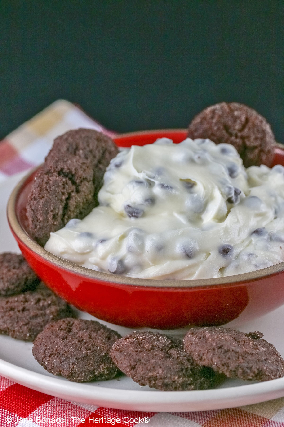 Chocolate Chip Toffee Cheesecake Cookie Dip is piled high in a red bowl on a white plate and surrounded with small chocolate cookies and red-skinned apple slices, all sitting on a red, white, and gold cloth © 2026 Jane Bonacci, The Heritage Cook.