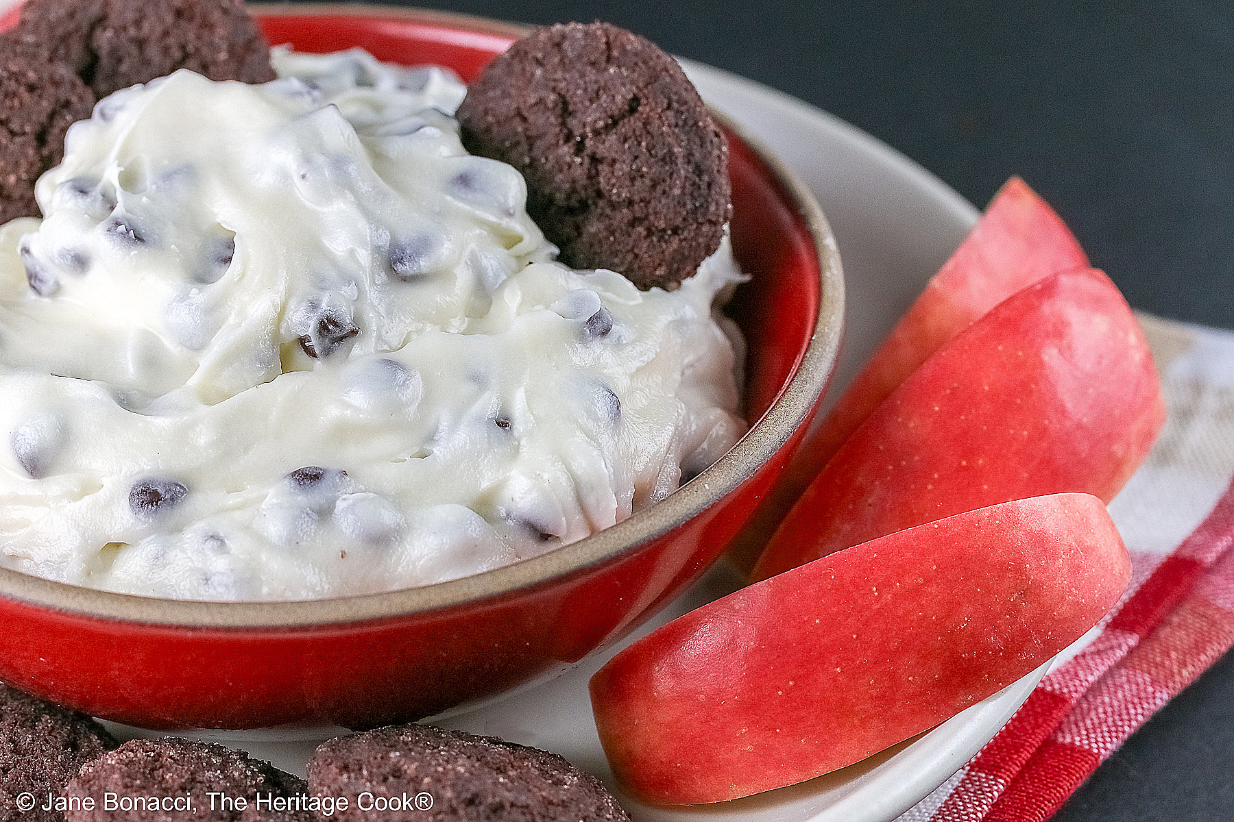 Chocolate Chip Toffee Cheesecake Cookie Dip is piled high in a red bowl on a white plate and surrounded with small chocolate cookies and red-skinned apple slices, all sitting on a red, white, and gold cloth © 2026 Jane Bonacci, The Heritage Cook.