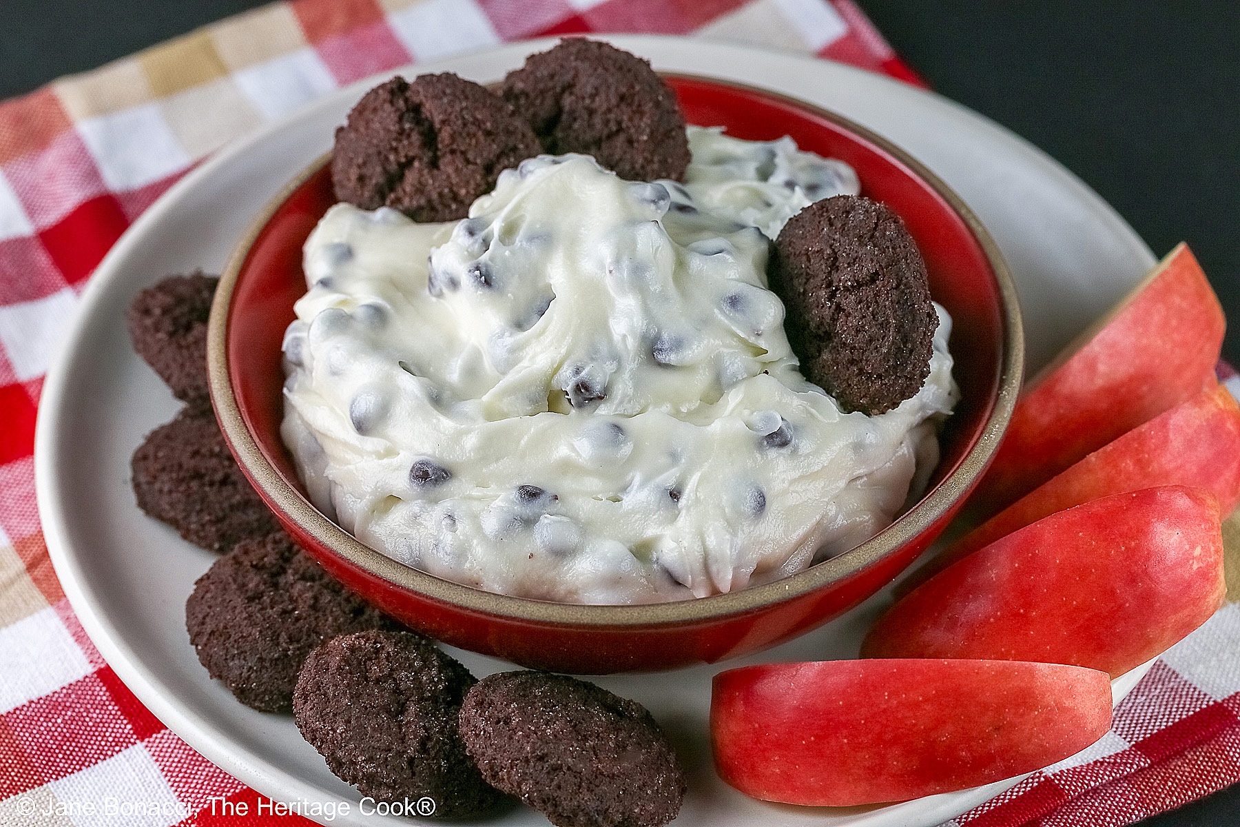 Chocolate Chip Toffee Cheesecake Cookie Dip is piled high in a red bowl on a white plate and surrounded with small chocolate cookies and red-skinned apple slices, all sitting on a red, white, and gold cloth © 2026 Jane Bonacci, The Heritage Cook.