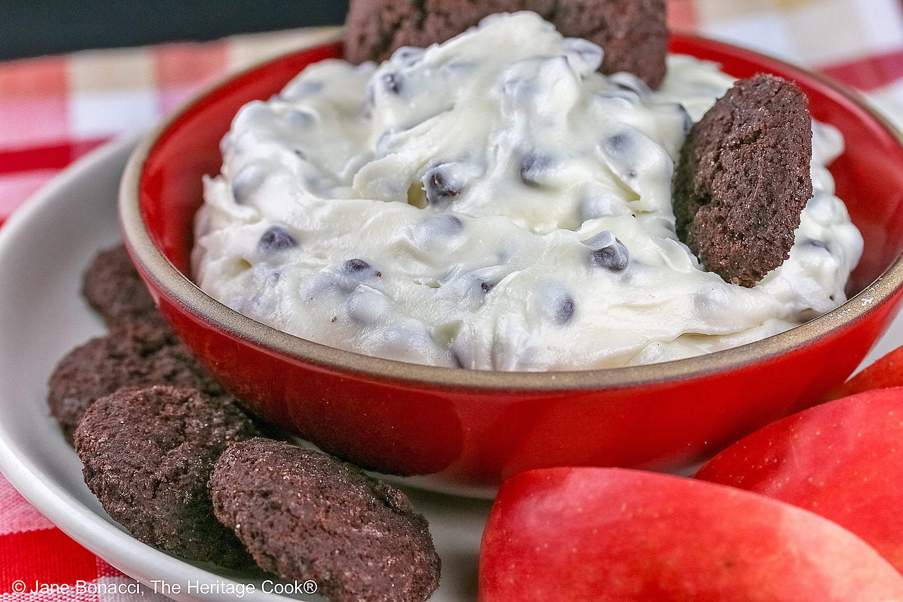 Chocolate Chip Toffee Cheesecake Cookie Dip is piled high in a red bowl on a white plate and surrounded with small chocolate cookies and red-skinned apple slices, all sitting on a red, white, and gold cloth © 2026 Jane Bonacci, The Heritage Cook.