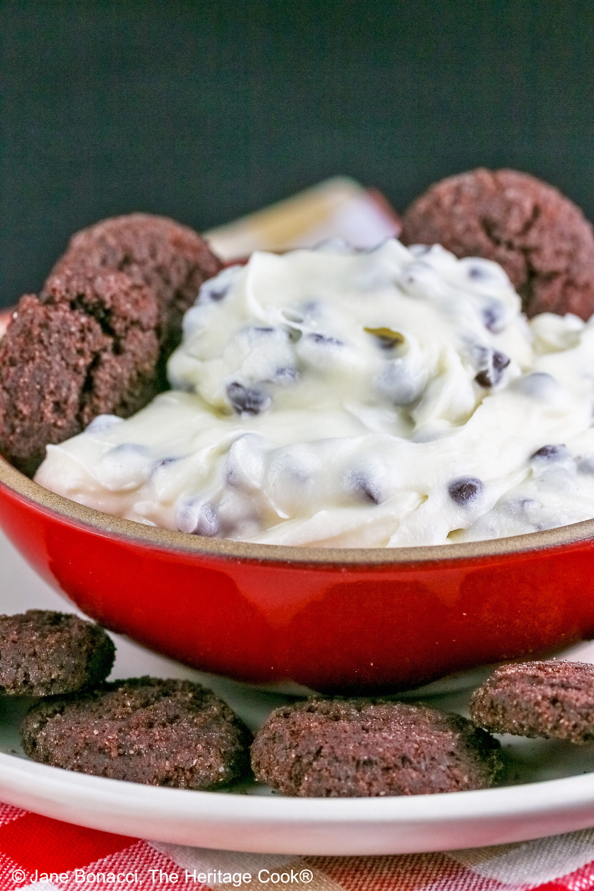 Chocolate Chip Toffee Cheesecake Cookie Dip is piled high in a red bowl on a white plate and surrounded with small chocolate cookies and red-skinned apple slices, all sitting on a red, white, and gold cloth © 2026 Jane Bonacci, The Heritage Cook.