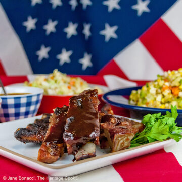 Melt in Your Mouth Ribs with a dry rub on them, wrapped tightly in foil, then cooked to beautiful tenderness, then served brushed with BBQ sauce on a white plate and served with coleslaw, baked beans, and corn salad sitting on an American flag in celebration of the 4th of July © 2026, Jane Bonacci, The Heritage Cook.