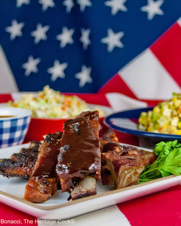 Melt in Your Mouth Ribs with a dry rub on them, wrapped tightly in foil, then cooked to beautiful tenderness, then served brushed with BBQ sauce on a white plate and served with coleslaw, baked beans, and corn salad sitting on an American flag in celebration of the 4th of July © 2026, Jane Bonacci, The Heritage Cook.