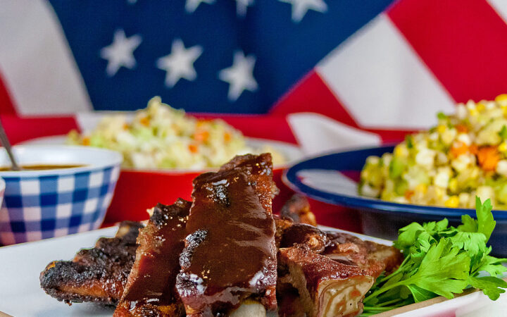 Melt in Your Mouth Ribs with a dry rub on them, wrapped tightly in foil, then cooked to beautiful tenderness, then served brushed with BBQ sauce on a white plate and served with coleslaw, baked beans, and corn salad sitting on an American flag in celebration of the 4th of July © 2026, Jane Bonacci, The Heritage Cook.