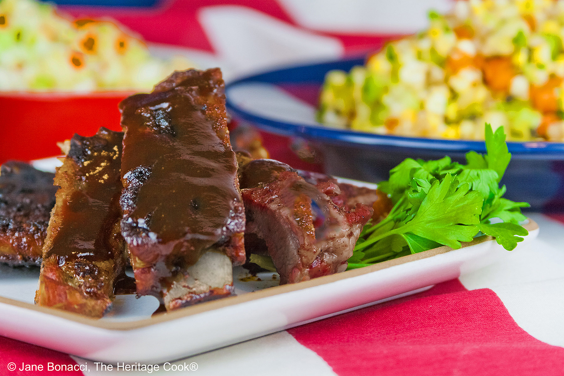 Melt in Your Mouth Ribs with a dry rub on them, wrapped tightly in foil, then cooked to beautiful tenderness, then served brushed with BBQ sauce on a white plate and served with coleslaw, baked beans, and corn salad sitting on an American flag in celebration of the 4th of July © 2026, Jane Bonacci, The Heritage Cook.