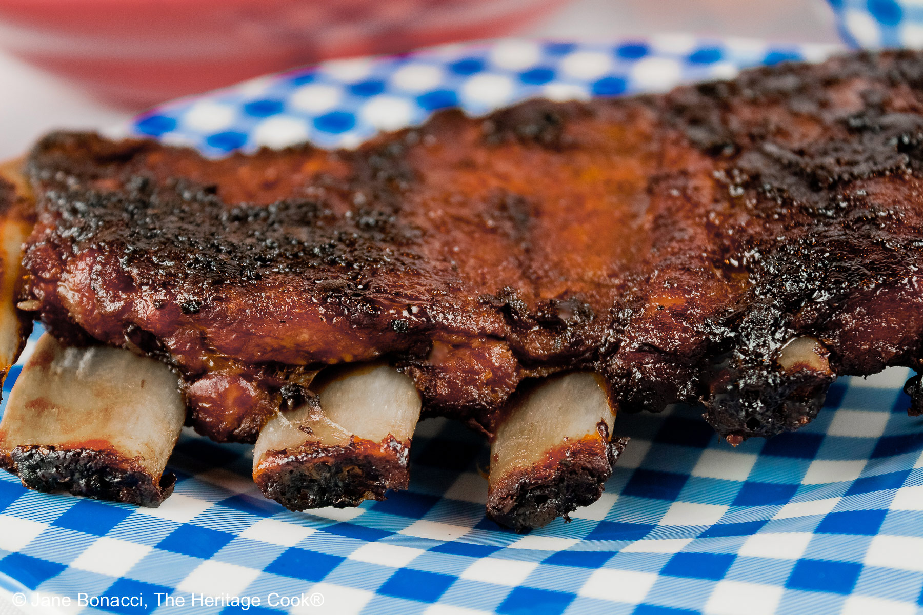 Melt in Your Mouth Ribs with a dry rub on them, wrapped tightly in foil, then cooked to beautiful tenderness, then served brushed with BBQ sauce on a white plate and served with coleslaw, baked beans, and corn salad sitting on an American flag in celebration of the 4th of July © 2026, Jane Bonacci, The Heritage Cook.