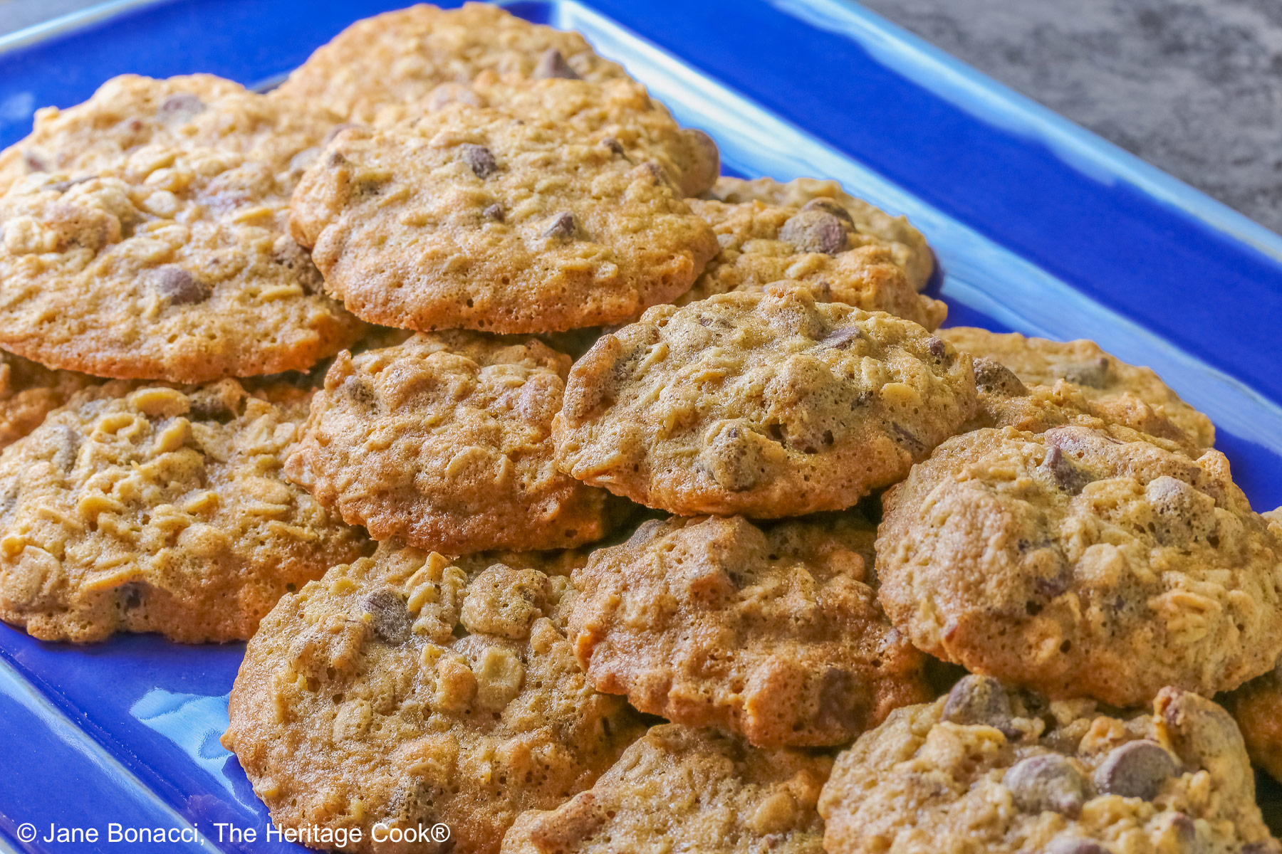 A large pile of amazing, delicious Chewy Oatmeal Chocolate Chip Cookies stacked on a rectangular blue platter sitting on a mottled gray background © 2026 Jane Bonacci, The Heritage Cook.
