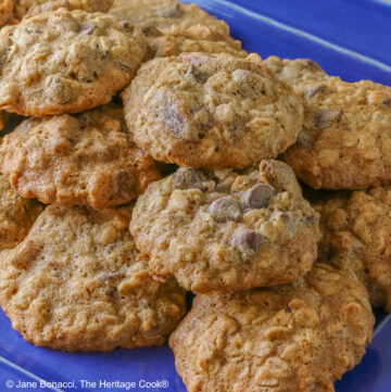A large pile of amazing, delicious Chewy Oatmeal Chocolate Chip Cookies stacked on a rectangular blue platter sitting on a mottled gray background © 2026 Jane Bonacci, The Heritage Cook.
